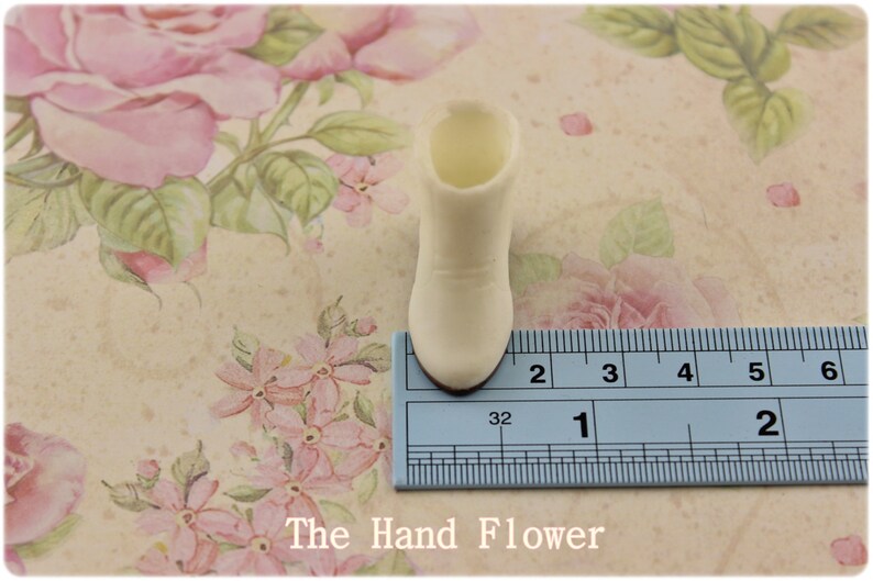 Puede incluir: Una peque&ntilde;a bota de color crema, de unos 3,2 cm de altura, se muestra sobre una superficie con estampado floral. La bota tiene una punta redondeada y un peque&ntilde;o tac&oacute;n. El fondo presenta un dise&ntilde;o floral vintage con rosas rosas y hojas verdes.