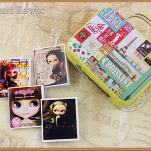 Puede incluir: Cuatro libros en miniatura con cubiertas coloridas que presentan ilustraciones de muñecas con ojos grandes. Los libros están dispuestos sobre un fondo beige con una maleta amarilla con temática parisina.