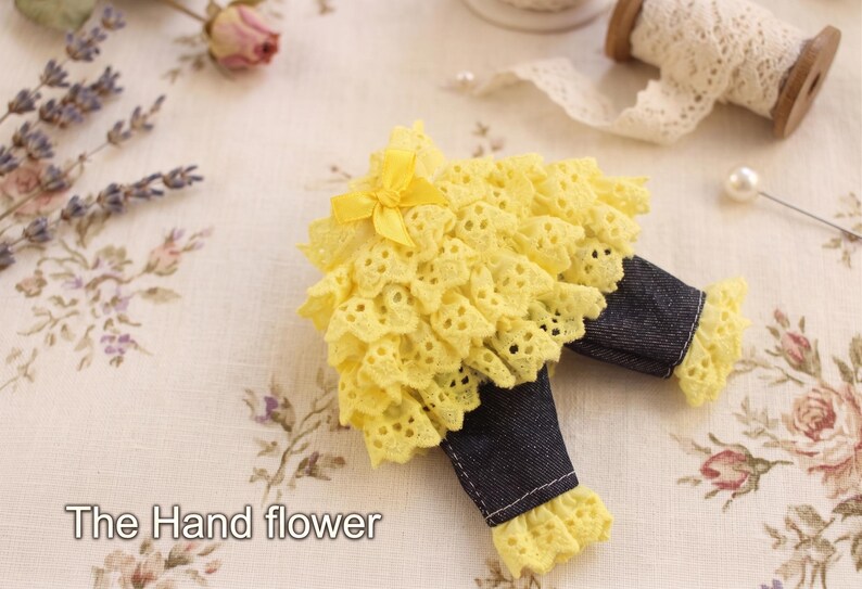 May include: A decorative item featuring a bright yellow eyelet lace skirt with a small matching yellow ribbon bow, paired with dark blue denim trousers that have yellow lace ruffles at the cuffs. The text The Hand flower is visible in the corner.