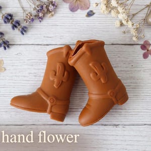 May include: A pair of brown cowboy boots featuring a raised cactus design on the shaft and a decorative strap detail across the ankle. The boots are positioned side by side. The text "The hand flower" is printed in white at the bottom left.