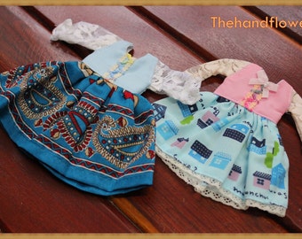 Puede incluir: Tres vestidos de muñeca en miniatura con mangas de encaje. Uno es turquesa con un estampado de cachemira, otro es azul claro y el tercero es azul claro con un estampado de casas. Los vestidos están sobre una superficie de madera.