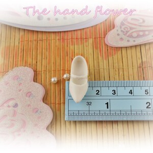 May include: A white ceramic shoe measuring 1 inch in length. The shoe is placed on a ruler for size reference.
