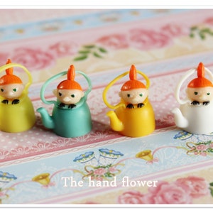 May include: Four small ceramic teapots with orange hair and painted faces. The teapots are yellow, green, white, and yellow. The teapots are on a pink and white striped surface.