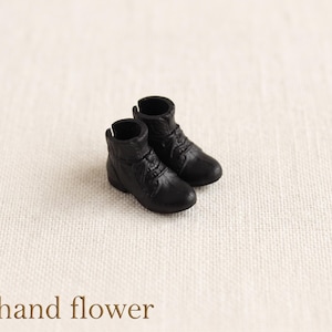 May include: A pair of miniature black boots with laces, set against a neutral background. The boots have a textured appearance and are designed to resemble real footwear. The text "The hand flower" is at the bottom of the image.
