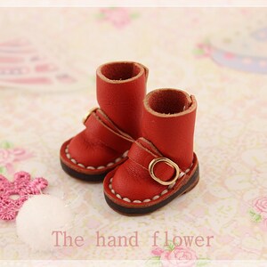 May include: A pair of miniature red leather boots with gold buckles and white stitching. The boots are small, with a short shaft and a rounded toe. The text "The hand flower" is visible at the bottom of the image.