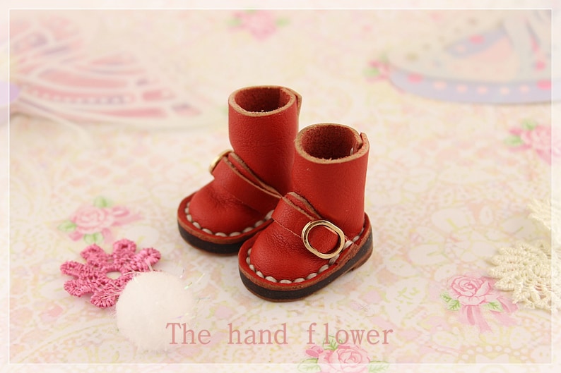 May include: A pair of miniature red leather boots with gold buckles and white stitching. The boots are small, with a short shaft and a rounded toe. The text "The hand flower" is visible at the bottom of the image.
