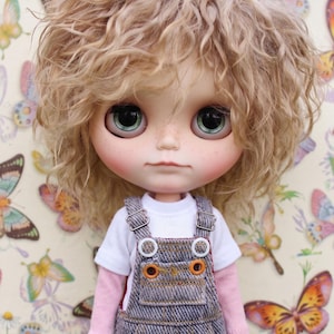 May include: A Blythe doll with light blonde curly hair and large green eyes. The doll wears a white t-shirt, a pink long-sleeved shirt, and denim overalls with orange stitching. The background features a butterfly pattern. The text "The Hand Flower" is visible.