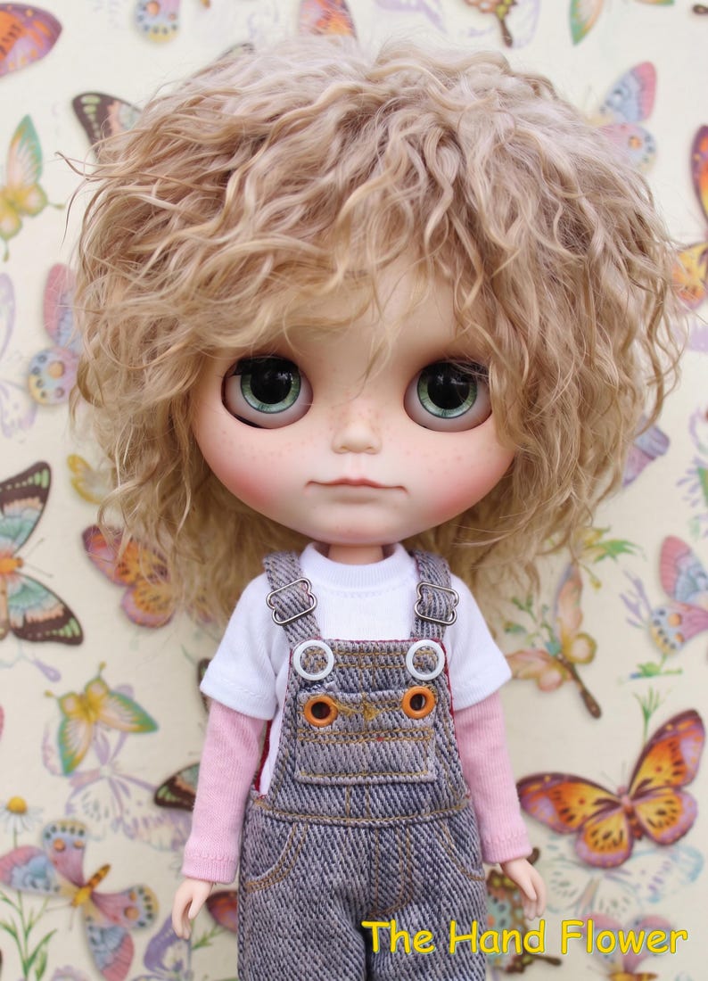 May include: A Blythe doll with light blonde curly hair and large green eyes. The doll wears a white t-shirt, a pink long-sleeved shirt, and denim overalls with orange stitching. The background features a butterfly pattern. The text "The Hand Flower" is visible.