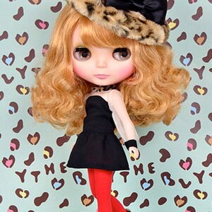 May include: A Blythe doll with wavy blonde hair wearing a leopard print faux fur hat with a black satin bow, a black sleeveless dress, bright red leggings, and black ankle boots with leopard print cuffs. The text 2013 Hasbro is visible.