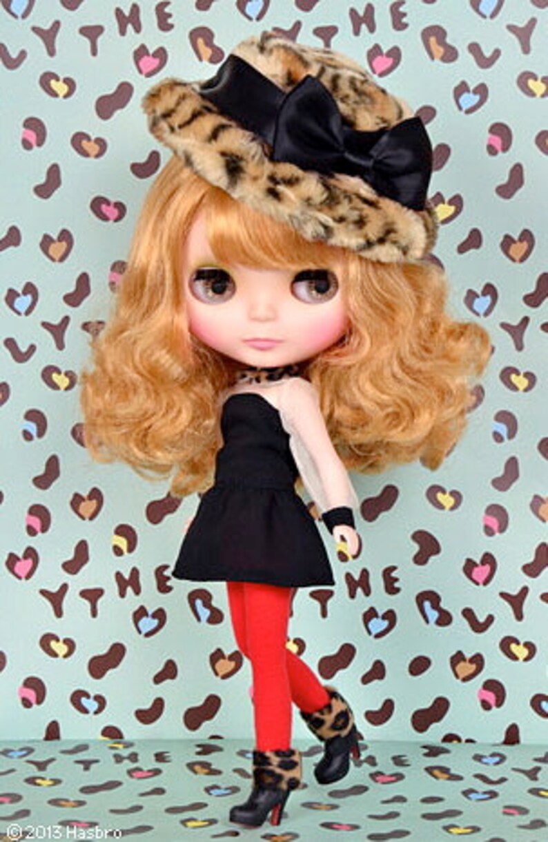May include: A Blythe doll with wavy blonde hair wearing a leopard print faux fur hat with a black satin bow, a black sleeveless dress, bright red leggings, and black ankle boots with leopard print cuffs. The text 2013 Hasbro is visible.