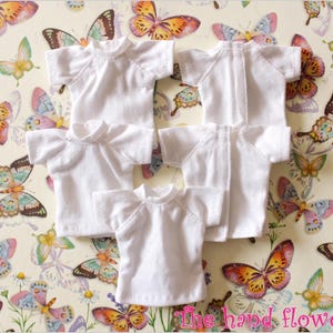 May include: Several small, white t-shirts with short sleeves and a crew neck are arranged on a patterned background. The background features a repeating design of colorful butterflies and flowers. The text "The hand flower" is visible in the bottom right corner.