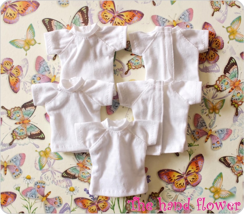 May include: Several small, white t-shirts with short sleeves and a crew neck are arranged on a patterned background. The background features a repeating design of colorful butterflies and flowers. The text "The hand flower" is visible in the bottom right corner.