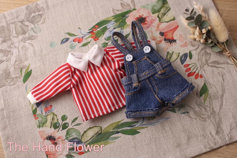 May include: A miniature outfit featuring a red and white striped shirt with a white collar and denim overalls. The outfit is displayed on a floral patterned fabric. The text "The Hand Flower" is visible at the bottom.