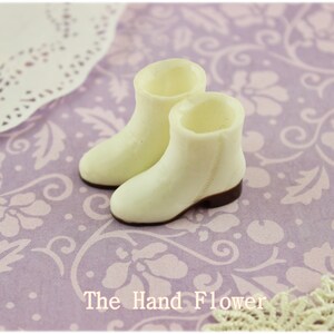 Puede incluir: Un par de botines en miniatura de color crema con suelas marrones. Est&aacute;n colocados sobre un fondo lavanda con un estampado floral. El texto "The Hand Flower" es visible en la parte inferior.
