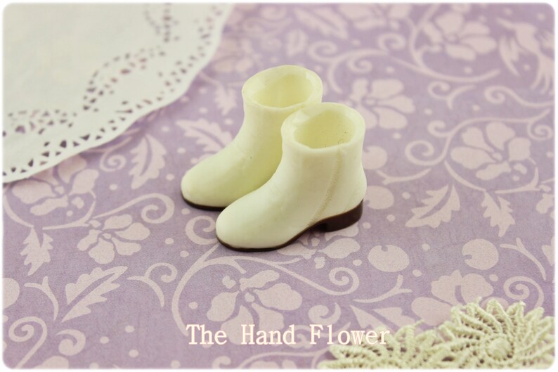 Puede incluir: Un par de botines en miniatura de color crema con suelas marrones. Est&aacute;n colocados sobre un fondo lavanda con un estampado floral. El texto "The Hand Flower" es visible en la parte inferior.