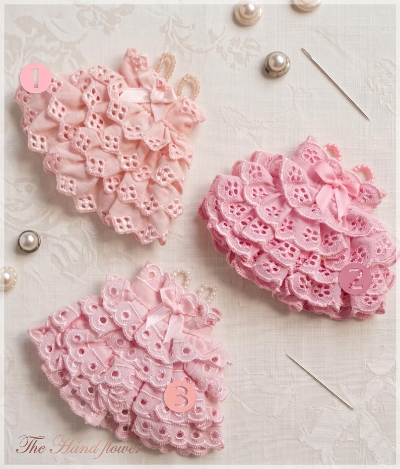 May include: Three pink fabric flower embellishments featuring layered eyelet lace, small satin bows, and pearl bead accents. Each piece is numbered one through three, accompanied by sewing needles and loose pearl beads on a textured surface.