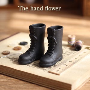 May include: A pair of miniature black lace-up boots with a matte finish, positioned next to a wooden ruler showing a scale in centimeters. The text "The hand flower" is visible above the boots, which feature molded eyelets and laces.