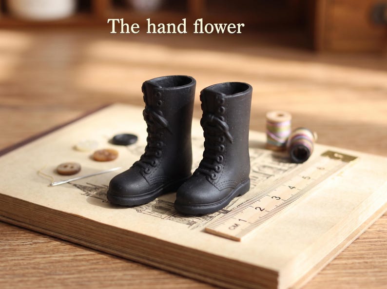 May include: A pair of miniature black lace-up boots with a matte finish, positioned next to a wooden ruler showing a scale in centimeters. The text "The hand flower" is visible above the boots, which feature molded eyelets and laces.