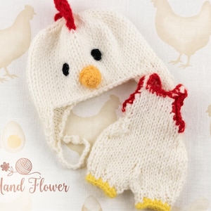 May include: A knitted white chicken hat and romper set with red and yellow accents. The hat features black eyes and an orange beak. The romper has red trim and yellow cuffs. The Hand Flower logo is in the bottom left corner.
