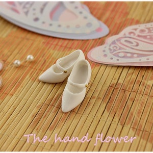 May include: Two white miniature shoes with gold buckles on a woven bamboo mat. The shoes are pointed toe and have a strap across the top.