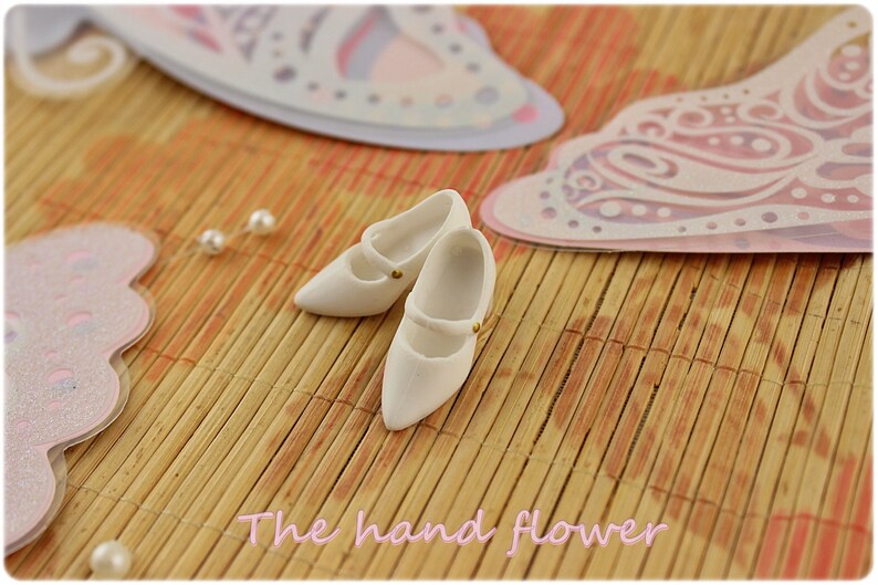 May include: Two white miniature shoes with gold buckles on a woven bamboo mat. The shoes are pointed toe and have a strap across the top.