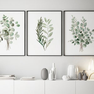 May include: Three framed watercolor prints of green eucalyptus leaves on a white wall above a white cabinet with a white ceramic vase and a gray ceramic vase on top.