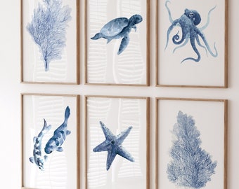 Puede incluir: Seis impresiones en acuarela enmarcadas con vida marina en tonos azules. Los temas incluyen coral, una tortuga, un pulpo, peces koi, una estrella de mar y algas marinas. Debajo, una unidad de almacenamiento blanca con formas recortadas y una pequeña mesa de madera.