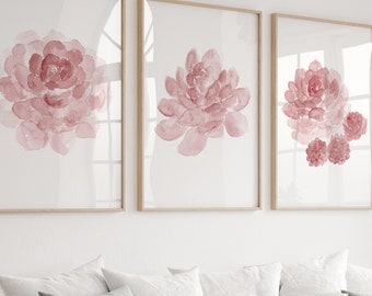 May include: Three framed watercolor paintings of pink flowers on a white wall above a white couch with pillows and a throw blanket. A vase of pink flowers sits on a wooden coffee table.