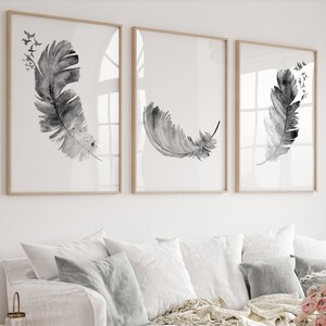 May include: Three framed black and white watercolor prints of feathers with a white background. The prints are hanging on a white wall above a white couch with gray pillows and a throw blanket. A vase of pink flowers sits on a wooden coffee table.