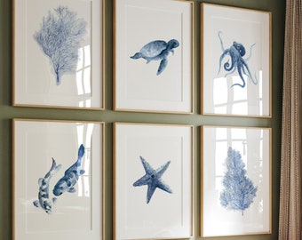 Puede incluir: Un conjunto de seis estampados acuarelados enmarcados que presentan ilustraciones azules y blancas de vida marina y coral. Las estampas se muestran en una pared en un ambiente de sala de estar.