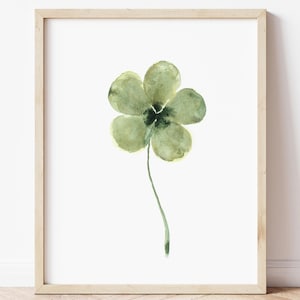 Op de afbeelding: Een aquarel van een groene klaver met vier bladeren op een witte achtergrond. Het schilderij is ingelijst in een licht houten lijst.