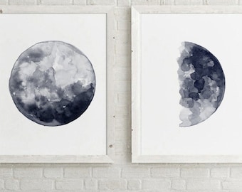 May include: Two framed watercolor paintings of the moon in different phases. The first painting shows a full moon in shades of gray. The second painting shows a half moon in shades of gray.