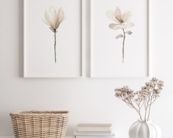 Könnte beinhalten: Zwei gerahmte Aquarelldrucke von Magnolienblüten in Braun- und Weißtönen, die an einer weißen Wand über einem Weidenkorb, einem Stapel Büchern und einer weißen Keramikvase mit Trockenblumen hängen.