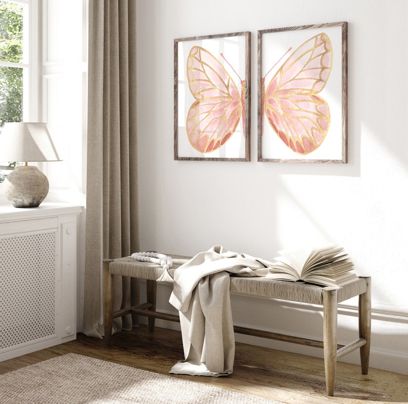 May include: Two framed prints of pink and gold butterflies on a white wall above a wooden bench with a white throw blanket and a book on top.