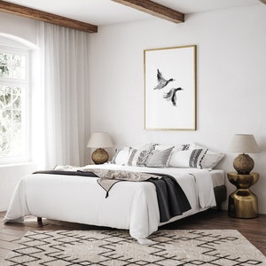 May include: A white bed with a black throw blanket and white pillows. The bed is in a bedroom with a large window, a wooden beam ceiling, and a framed print of two birds in flight.