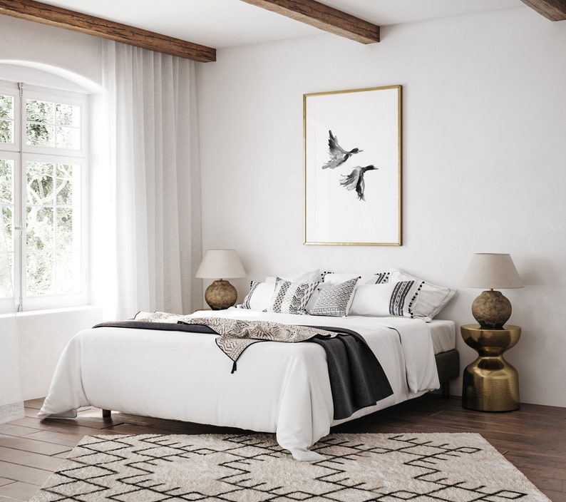 May include: A white bed with a black throw blanket and white pillows. The bed is in a bedroom with a large window, a wooden beam ceiling, and a framed print of two birds in flight.