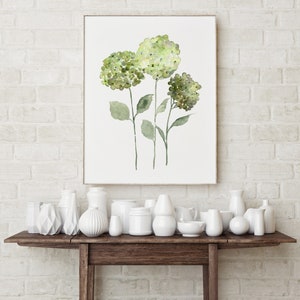 May include: A watercolor painting of three green hydrangea flowers on a white background. The painting is framed and hanging on a white brick wall above a wooden table with white ceramic vases.