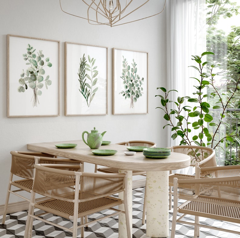 May include: A light wood dining table with a white and green tiled floor. The table has a light wood oval top and a white base. There are four chairs around the table, each with a light wood frame and a woven seat. There are three framed prints on the wall behind the table, each featuring a watercolor illustration of green leaves.