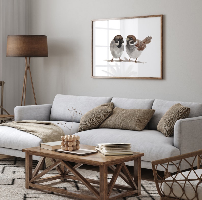 May include: A watercolor painting of two brown sparrows perched on a branch. The painting is framed in a light wood frame and hangs on a white wall above a gray couch with throw pillows.