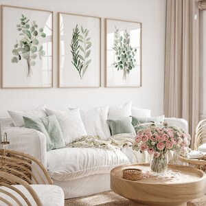 May include: Three framed watercolor prints of green leafy branches, hanging on a white wall above a white couch with green throw pillows and a wooden coffee table with a vase of pink flowers.