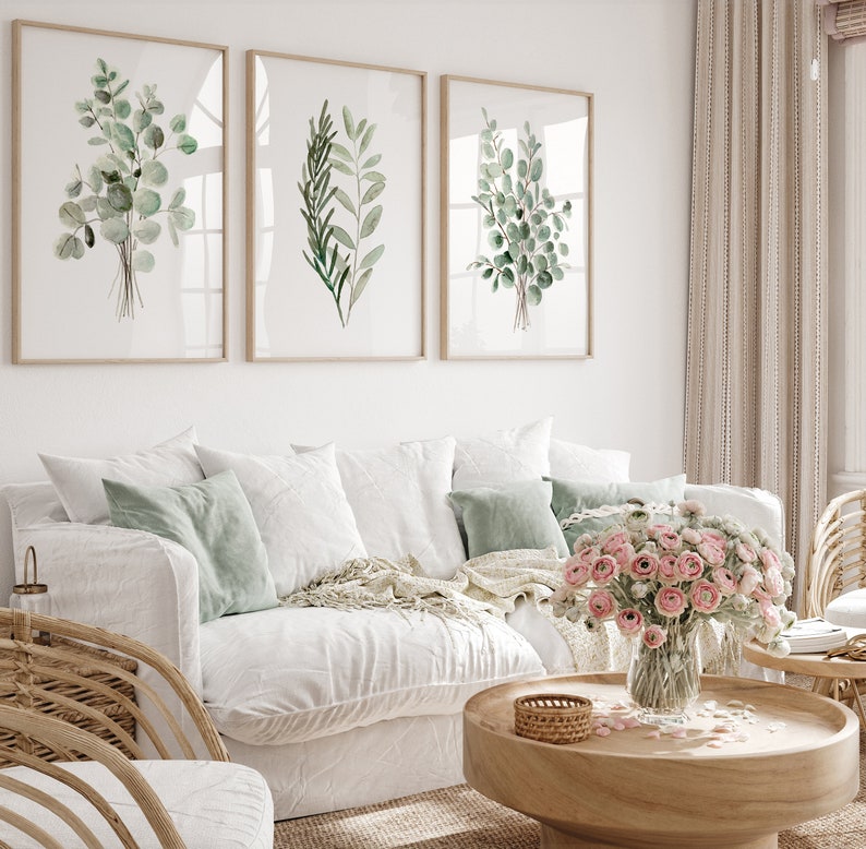 May include: Three framed watercolor prints of green leafy branches, hanging on a white wall above a white couch with green throw pillows and a wooden coffee table with a vase of pink flowers.