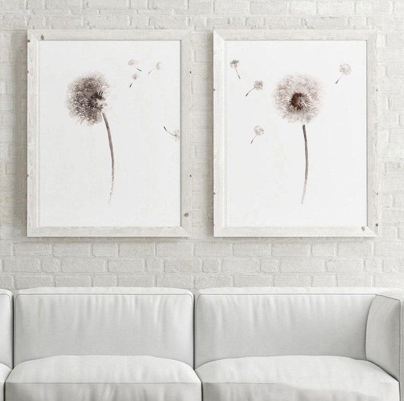 May include: Two framed watercolor paintings of dandelion flowers with seeds blowing in the wind. The paintings are in white frames and are hanging on a white brick wall.