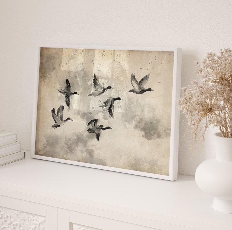May include: A framed art print featuring a flock of ducks in flight. The watercolor painting uses shades of gray and black against a beige background. The artwork is displayed in a white frame, adding a touch of elegance to the nature-inspired scene.