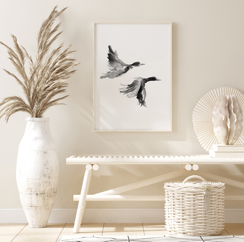 May include: A black and white watercolor print of two birds in flight, framed and hanging on a white wall above a white bench and a white wicker basket.