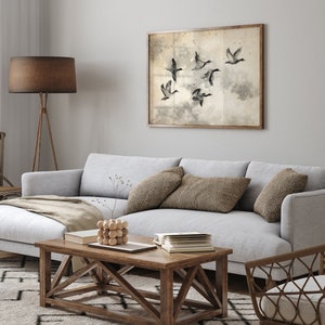 May include: A framed artwork featuring a flock of flying ducks in a sepia-toned watercolor style hangs above a light gray sofa. The living room setting includes a wooden coffee table, throw pillows, and a floor lamp with a brown shade.