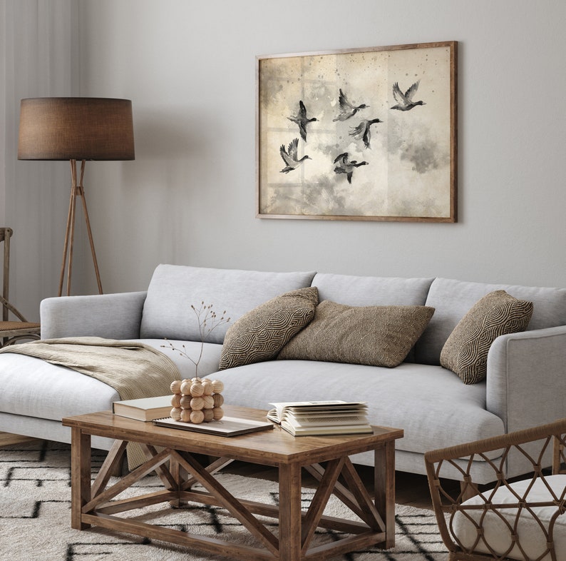 May include: A framed artwork featuring a flock of flying ducks in a sepia-toned watercolor style hangs above a light gray sofa. The living room setting includes a wooden coffee table, throw pillows, and a floor lamp with a brown shade.