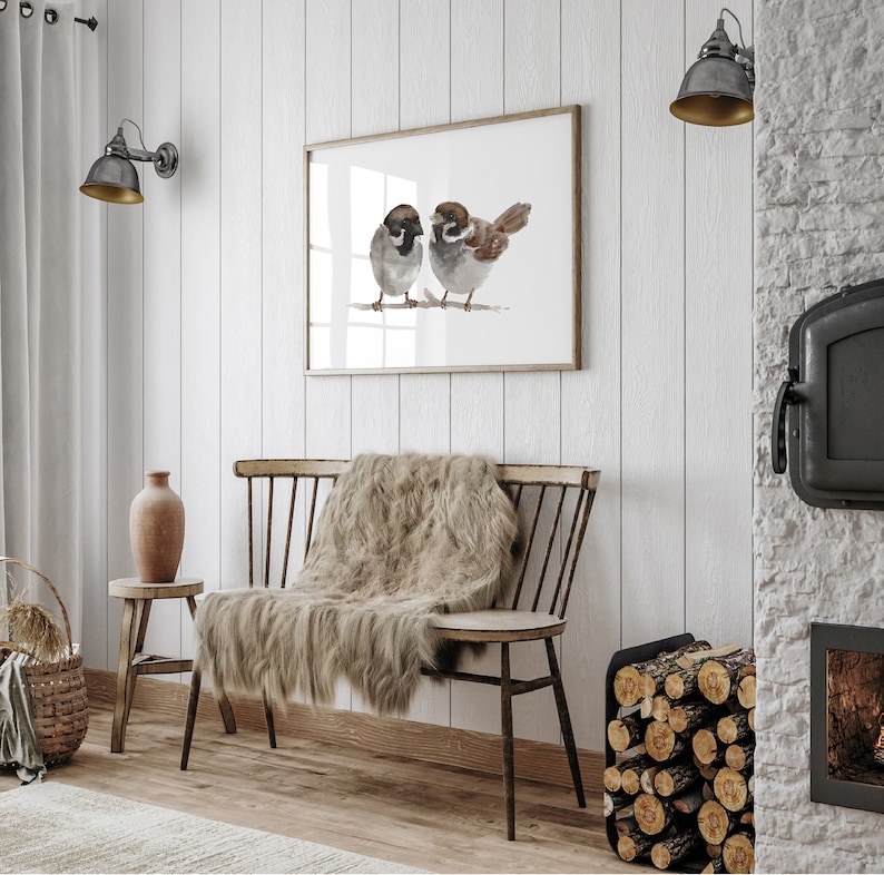 May include: A watercolor painting of two sparrows perched on a branch. The painting is framed and hanging on a white wall. The wall is made of wood planks. There is a wooden bench with a fur throw on it in front of the painting.