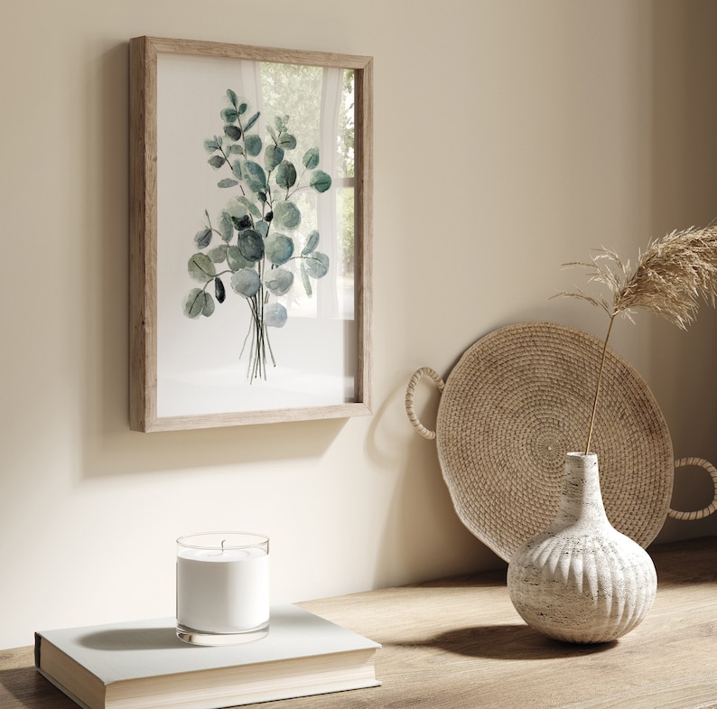 May include: A framed watercolor print of a bouquet of eucalyptus leaves in shades of green, displayed on a light wood shelf with a woven tray and a white ceramic vase with a dried grass stem.