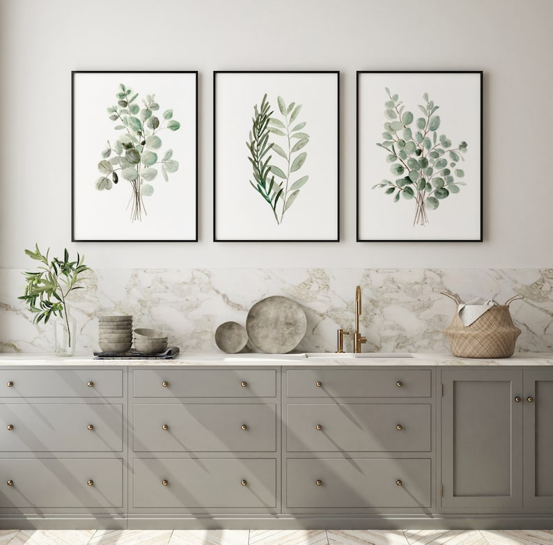 May include: Three framed watercolor prints of green foliage, hanging on a white wall above a gray kitchen cabinet with drawers and doors.
