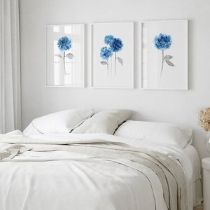 May include: Three watercolor paintings of blue hydrangeas on white backgrounds, displayed in a row on a white wall above a white bed with a light gray blanket.